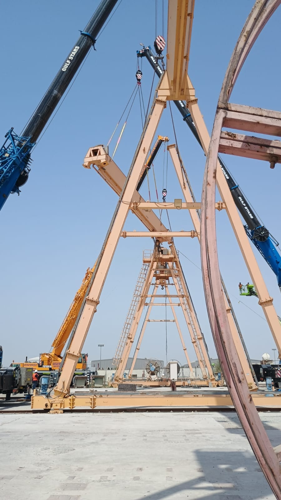 Looking up at a gantry crane part-assembled, its legs and cross members held by mobile cranes while an access platform reaches the joint