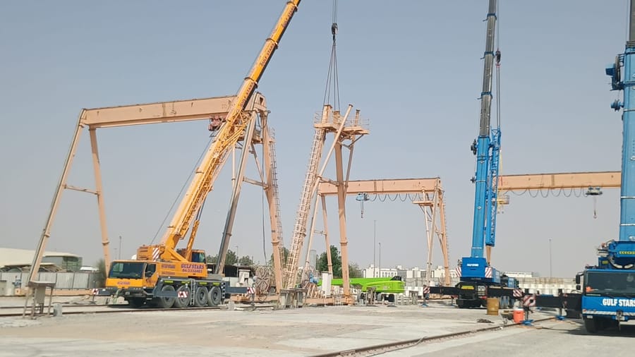 Gantry crane erection in an open yard, with two mobile cranes supporting the girder and legs and rail track laid across the ground