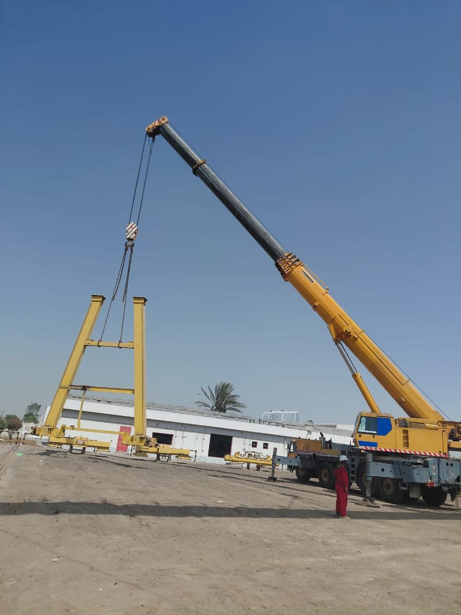 A yellow gantry crane frame suspended on chains from a mobile crane, being lowered into position in an open yard while a worker watches from the ground