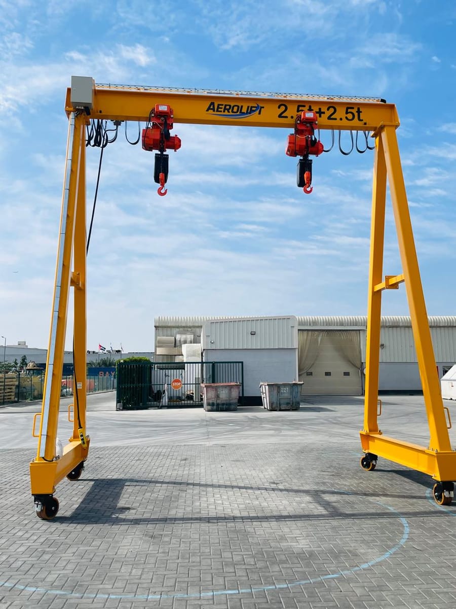 Aerolift-branded portable gantry crane marked 2.5 t plus 2.5 t, carrying two red electric chain hoists on the beam, standing on castors in an open yard