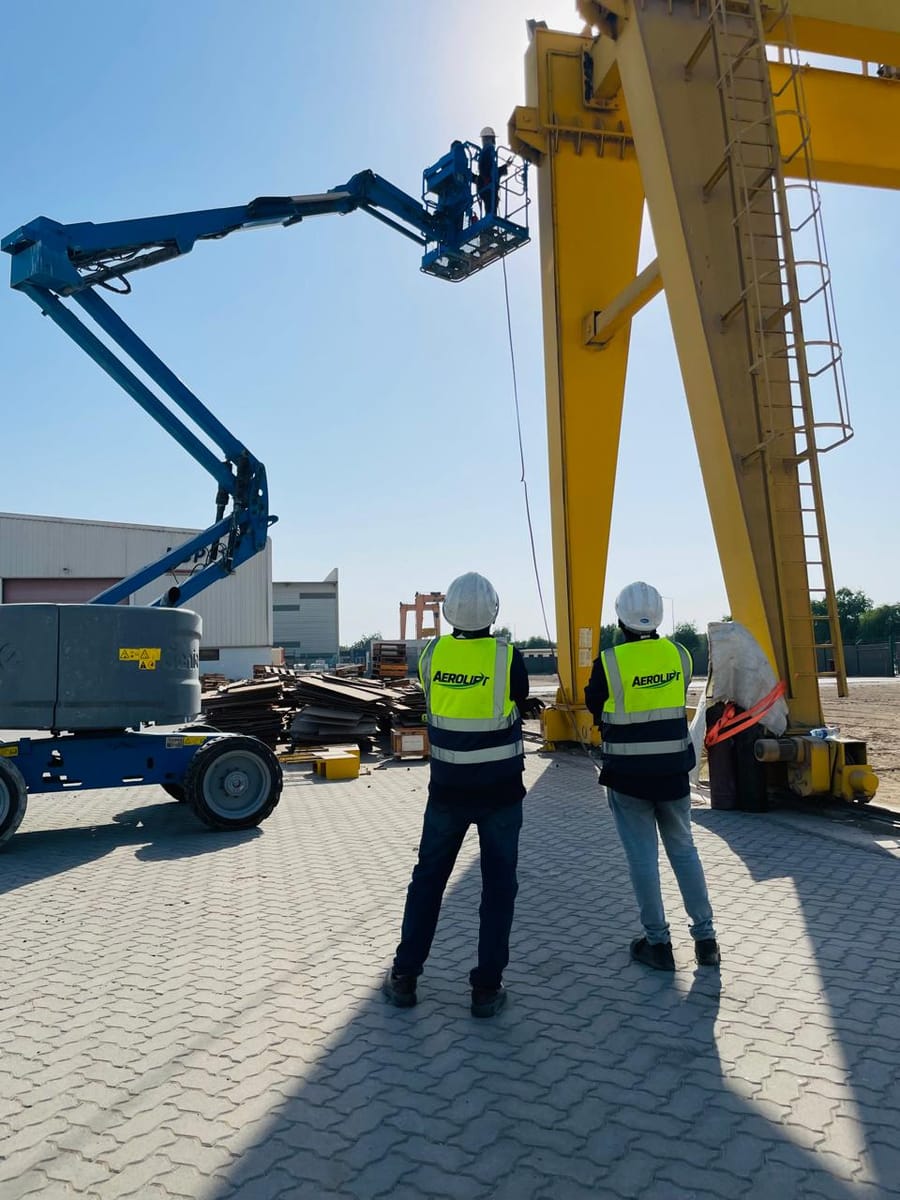 Two Aerolift technicians in branded hi-vis vests and hard hats watching work on the leg of a large yellow gantry crane, with a boom lift alongside